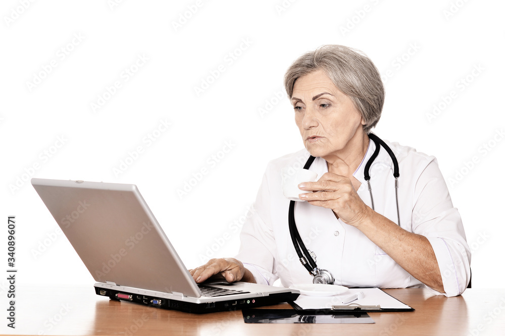 Serious elderly woman doctor sitting at table with computer on white Serious elderly woman doctor sitting at table with computer on white