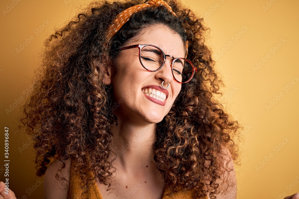 Beautiful brunette woman on vacation with curly hair and piercing wearing glasses and diadem very happy and excited doing winner gesture with arms raised, smiling and screaming for success Beautiful brunette woman on vacation with curly hair and piercing wearing glasses and diadem very happy and excited doing winner gesture with arms raised, smiling and screaming for success