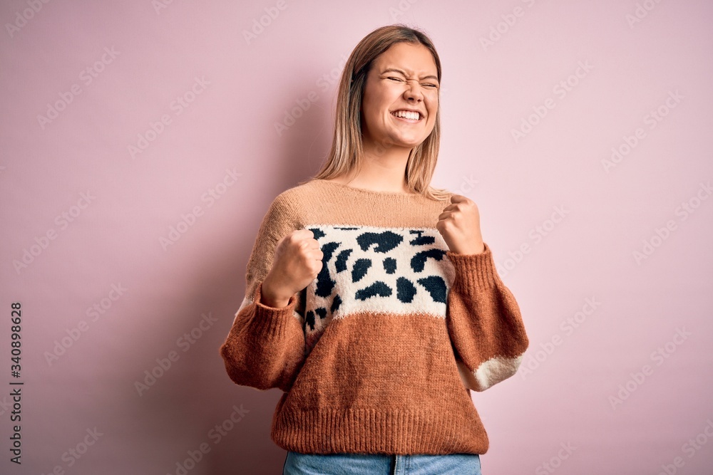 Young beautiful blonde woman wearing fashion animal print sweater over pink background very happy and excited doing winner gesture with arms raised, smiling and screaming for success. Celebration Young beautiful blonde woman wearing fashion animal print sweater over pink background very happy and excited doing winner gesture with arms raised, smiling and screaming for success. Celebration