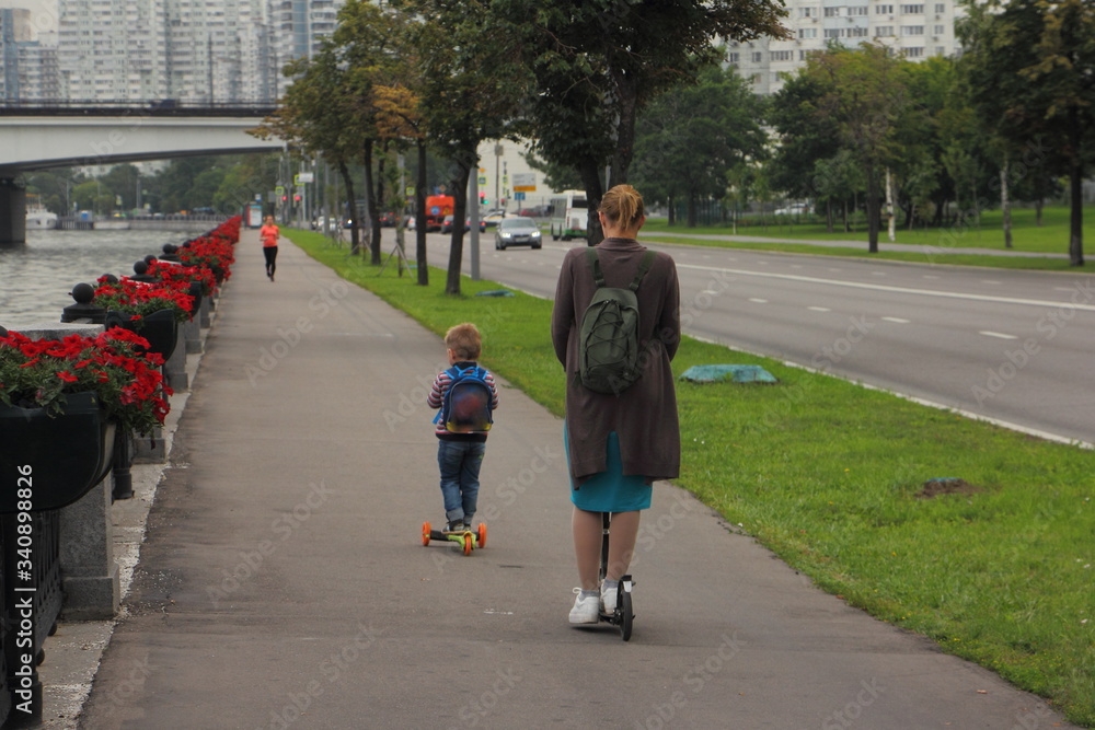 A woman and a child boy riding a kick scooters on a road in the Park at summer evening, outdoor vacation in big city