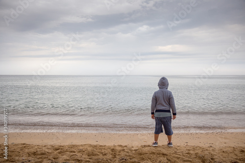 Fotografie Lonely hooded boy turned on his back looking out to sea on a gray and cloudy day