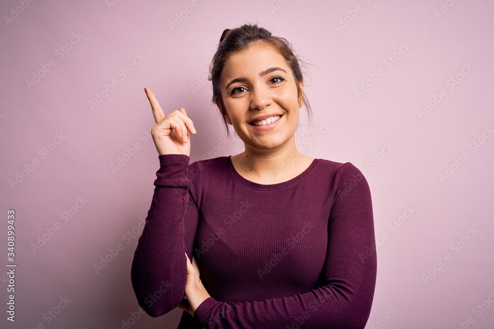 Beautiful young woman wearing casual bun hairstyle over pink isolated background with a big smile on face, pointing with hand and finger to the side looking at the camera. Beautiful young woman wearing casual bun hairstyle over pink isolated background with a big smile on face, pointing with hand and finger to the side looking at the camera.