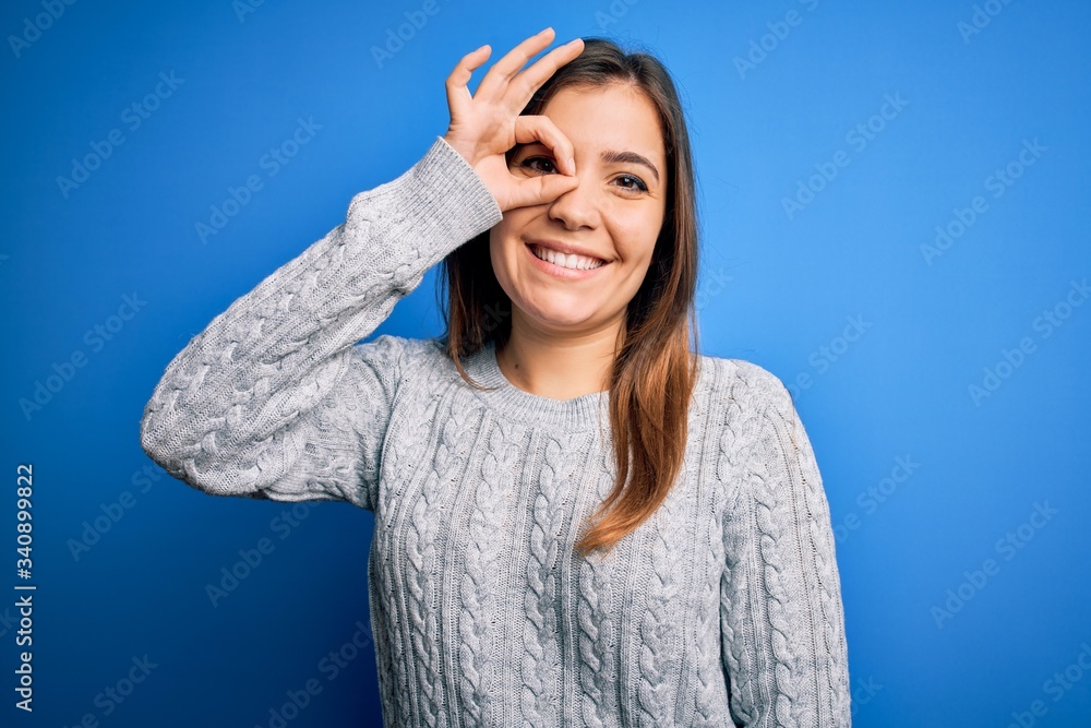Beautiful young woman wearing casual wool sweater standing over blue isolated background doing ok gesture with hand smiling, eye looking through fingers with happy face. Beautiful young woman wearing casual wool sweater standing over blue isolated background doing ok gesture with hand smiling, eye looking through fingers with happy face.