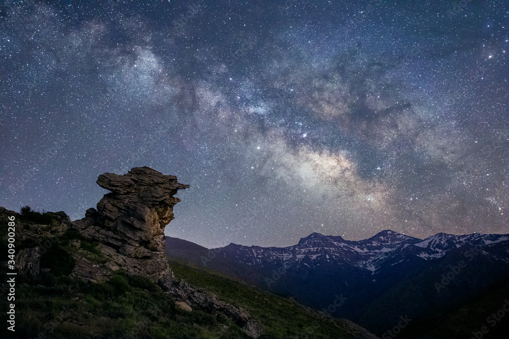 Panoramic night view of the Sierra Nevada peaks, Granada, Andalusia, with the Milky Way in the sky. Rocky landmark in the foreground and snowy mountains in the background.