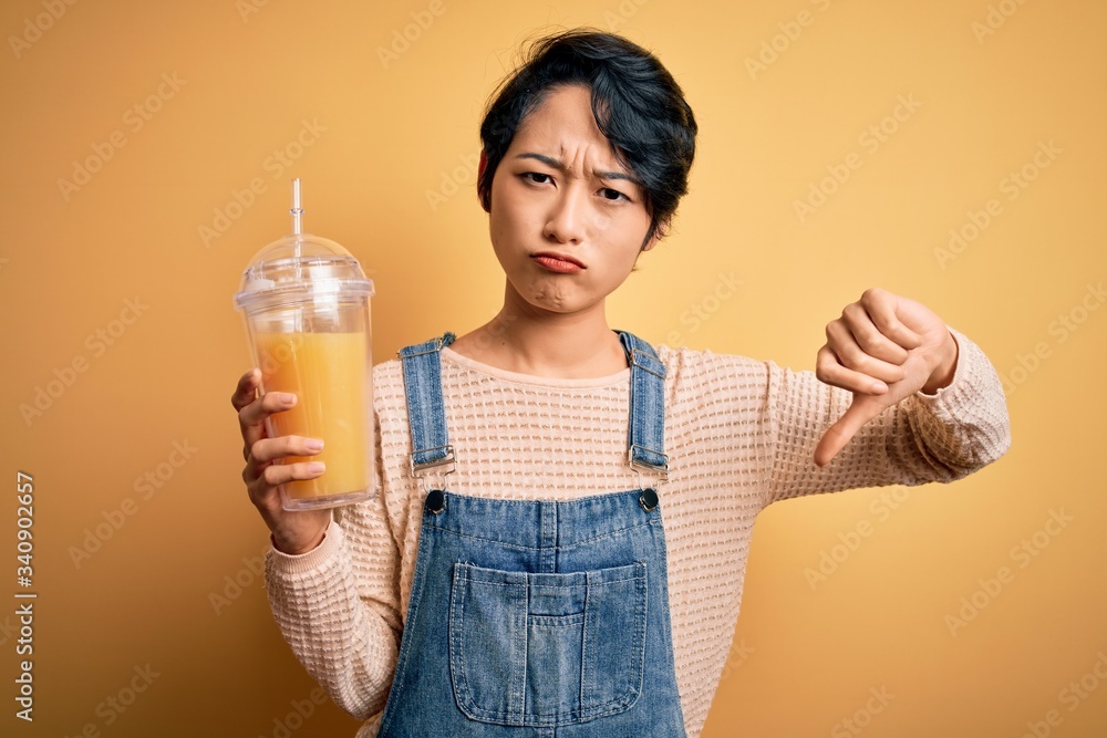Young beautiful chinese woman drinking healthy orange juice over isolated yellow background with angry face, negative sign showing dislike with thumbs down, rejection concept Young beautiful chinese woman drinking healthy orange juice over isolated yellow background with angry face, negative sign showing dislike with thumbs down, rejection concept