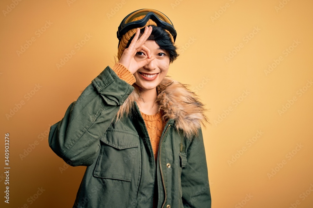 Beautiful asian skier girl wearing snow sportswear using ski goggles over yellow background doing ok gesture with hand smiling, eye looking through fingers with happy face.