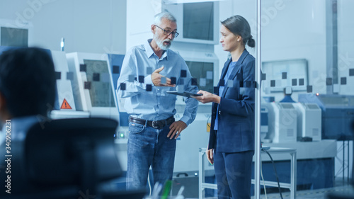 Modern Factory: Female Engineer, Male Project Manager Standing in High Tech Development Facility, Talking and Using Tablet Computer. Contemporary Facility with CNC Machinery, Robot Arm Production Line