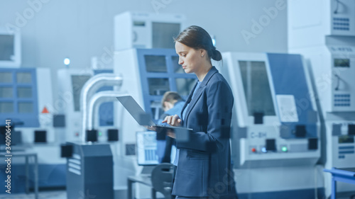  Modern Factory: Confident Female Engineer Walks Through Facility, Holding Laptop, Inspecting and Monitoring Workshop Workings. Manufactory Has Professionals Working on CNC Machinery, Robot Arm