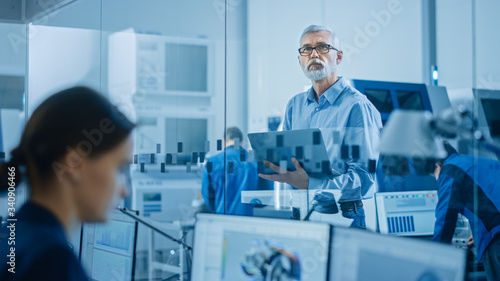Modern Factory: Senior Engineer Looks at Camera, Holding Laptop, Inspecting, Overlooking Workshop Operation. Manufactory Has Professionals Working on CNC Machinery, Robot Arm.