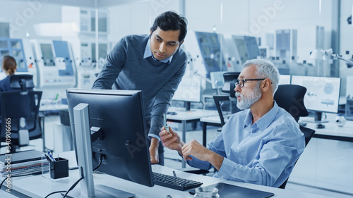 Modern Factory Office: Indian Project Manager Talks to Caucasian Senior Engineer whos Working on Computer, Inspect Metal Component. Professionals Solving Heavy Industry Problems, Having Discussion 