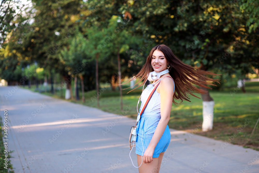Portrait of beautiful fashionista woman with long hair wearing blue romper and headphones outside in the park. 