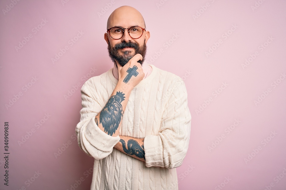 Handsome bald man with beard and tattoo wearing glasses and sweater over pink background looking confident at the camera smiling with crossed arms and hand raised on chin. Thinking positive. Handsome bald man with beard and tattoo wearing glasses and sweater over pink background looking confident at the camera smiling with crossed arms and hand raised on chin. Thinking positive.