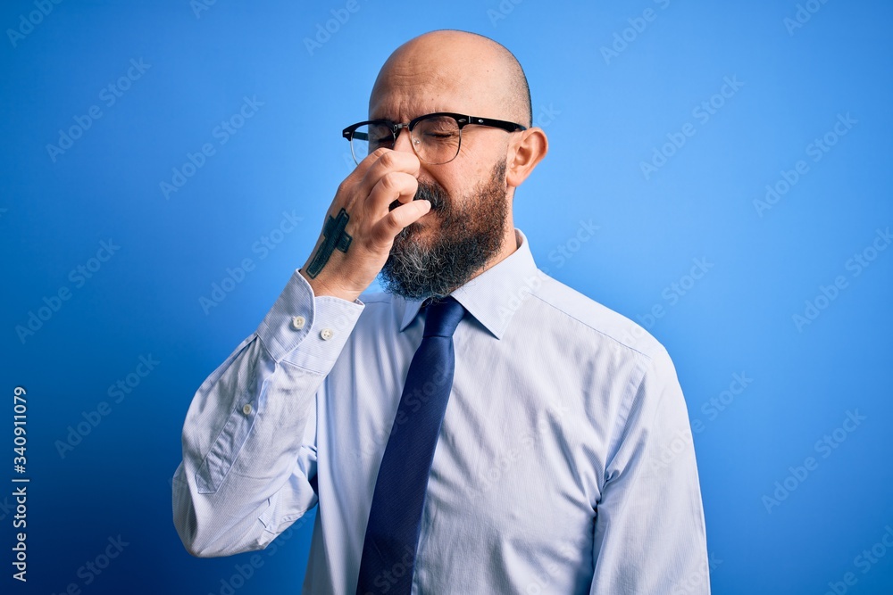 Handsome business bald man with beard wearing elegant tie and glasses over blue background smelling something stinky and disgusting, intolerable smell, holding breath with fingers on nose. Bad smell Handsome business bald man with beard wearing elegant tie and glasses over blue background smelling something stinky and disgusting, intolerable smell, holding breath with fingers on nose. Bad smell