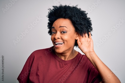 Young beautiful African American afro woman with curly hair wearing casual t-shirt standing smiling with hand over ear listening an hearing to rumor or gossip. Deafness concept.