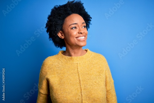 Young beautiful African American afro woman with curly hair wearing yellow casual sweater looking away to side with smile on face, natural expression. Laughing confident.