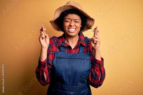 Young African American afro farmer woman with curly hair wearing apron and hat gesturing finger crossed smiling with hope and eyes closed. Luck and superstitious concept.