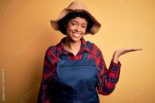 Young African American afro farmer woman with curly hair wearing apron and hat smiling cheerful presenting and pointing with palm of hand looking at the camera.