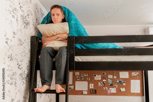 The guy is sitting on a bunk bed under a blue blanket and hugs a pillow, surprised, sad