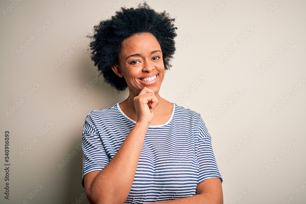 Young beautiful African American afro woman with curly hair wearing striped t-shirt looking confident at the camera with smile with crossed arms and hand raised on chin. Thinking positive. Young beautiful African American afro woman with curly hair wearing striped t-shirt looking confident at the camera with smile with crossed arms and hand raised on chin. Thinking positive.
