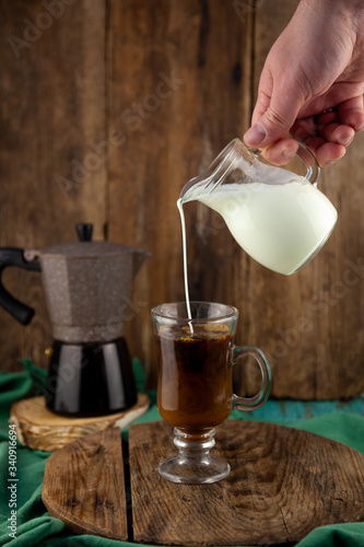 Milk is poured into decanter coffee. Geyser coffee maker, creamer on a wooden table. Glass cup with coffee making cappuccino. Making coffee, hand close-up.