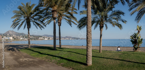 Malaga Beach La Malagueta wide panorama with palm trees and grass with people relaxing and sunbathing on the sand near the sea.