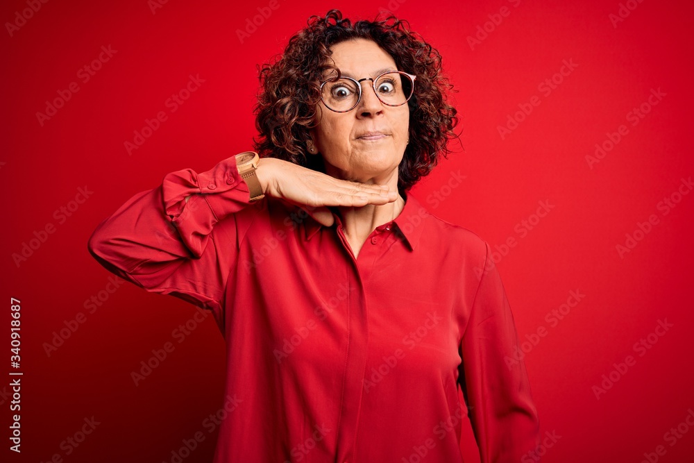 Middle age beautiful curly hair woman wearing casual shirt and glasses over red background cutting throat with hand as knife, threaten aggression with furious violence