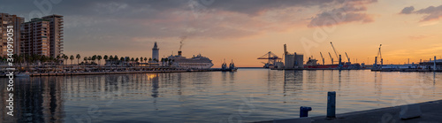 Sunset in Malaga Port wide panorama with purple cloudy sky. View of Quay 1 with palm trees and promenade, with cranes and a cruise ship sailing.