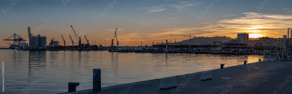 Sunset on the Port of Malaga Spain wide panorama. Docks with cranes silhouette with the sun setting behind the hills in backlight.