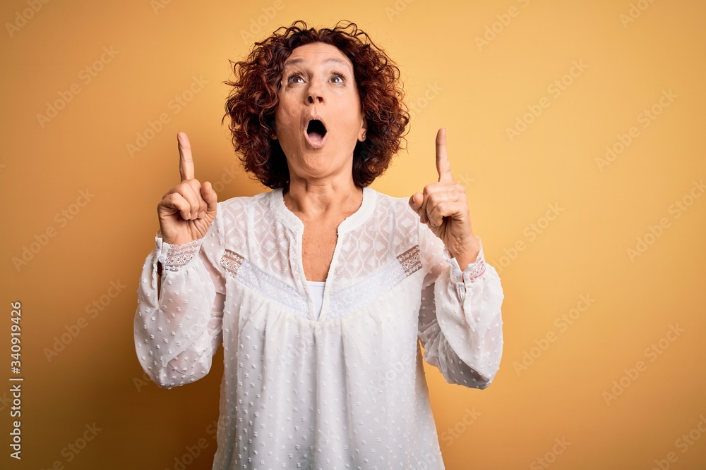 Middle age beautiful curly hair woman wearing casual summer dress over yellow background amazed and surprised looking up and pointing with fingers and raised arms. Middle age beautiful curly hair woman wearing casual summer dress over yellow background amazed and surprised looking up and pointing with fingers and raised arms.