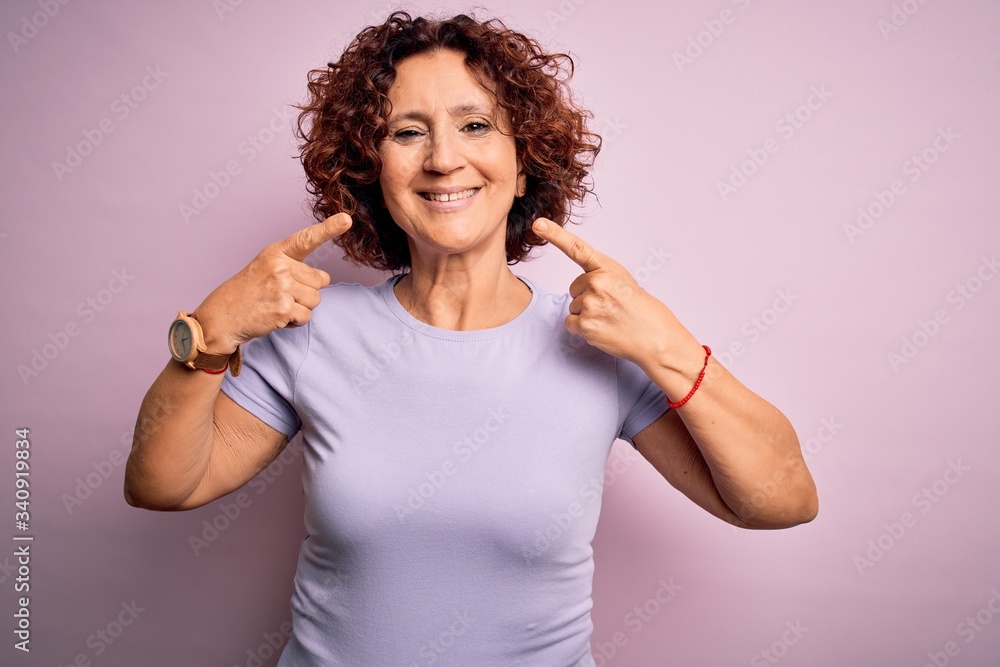Middle age beautiful curly hair woman wearing casual t-shirt over isolated pink background smiling cheerful showing and pointing with fingers teeth and mouth. Dental health concept. Middle age beautiful curly hair woman wearing casual t-shirt over isolated pink background smiling cheerful showing and pointing with fingers teeth and mouth. Dental health concept.