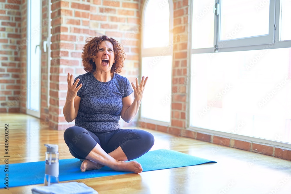 Middle age beautiful sportswoman wearing sportswear sitting on mat  practicing yoga at home crazy and mad shouting and yelling with aggressive  expression and arms raised. Frustration concept. Stock Photo