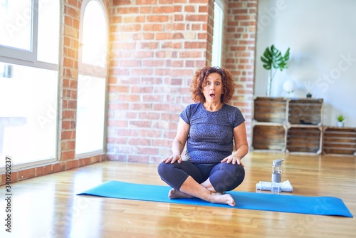 Middle age beautiful sportswoman wearing sportswear sitting on mat practicing yoga at home afraid and shocked with surprise expression, fear and excited face.