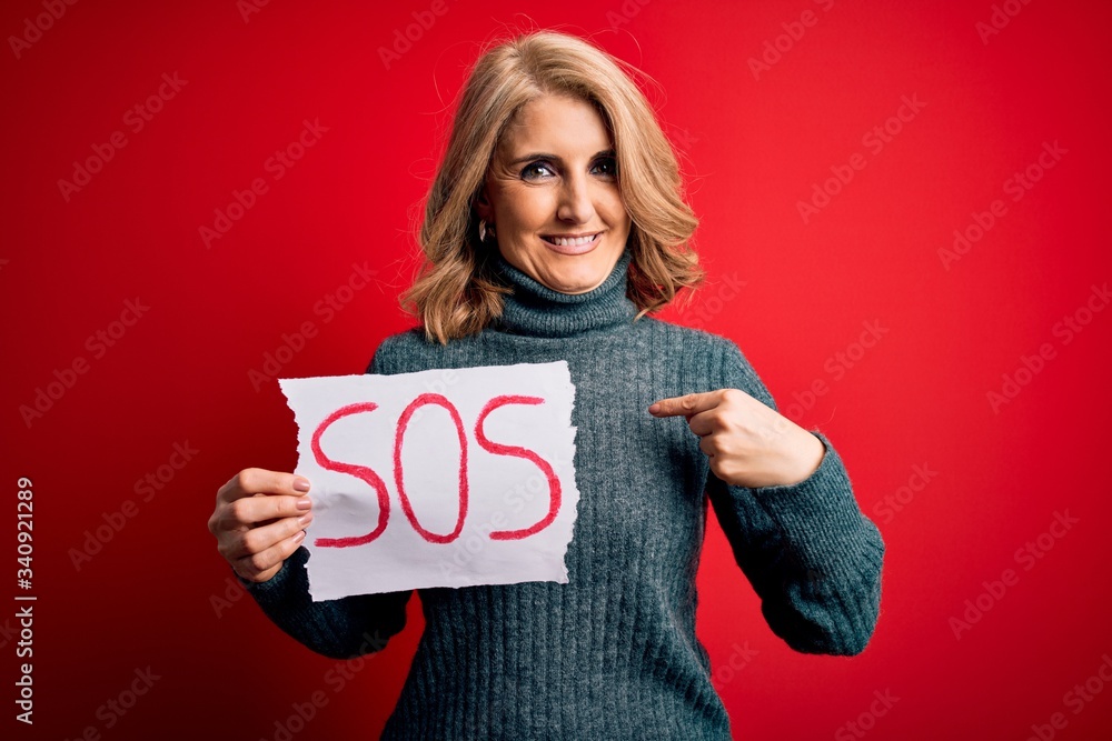 Middle age beautiful blonde woman holding paper with sos message over red bakground with surprise face pointing finger to himself Middle age beautiful blonde woman holding paper with sos message over red bakground with surprise face pointing finger to himself