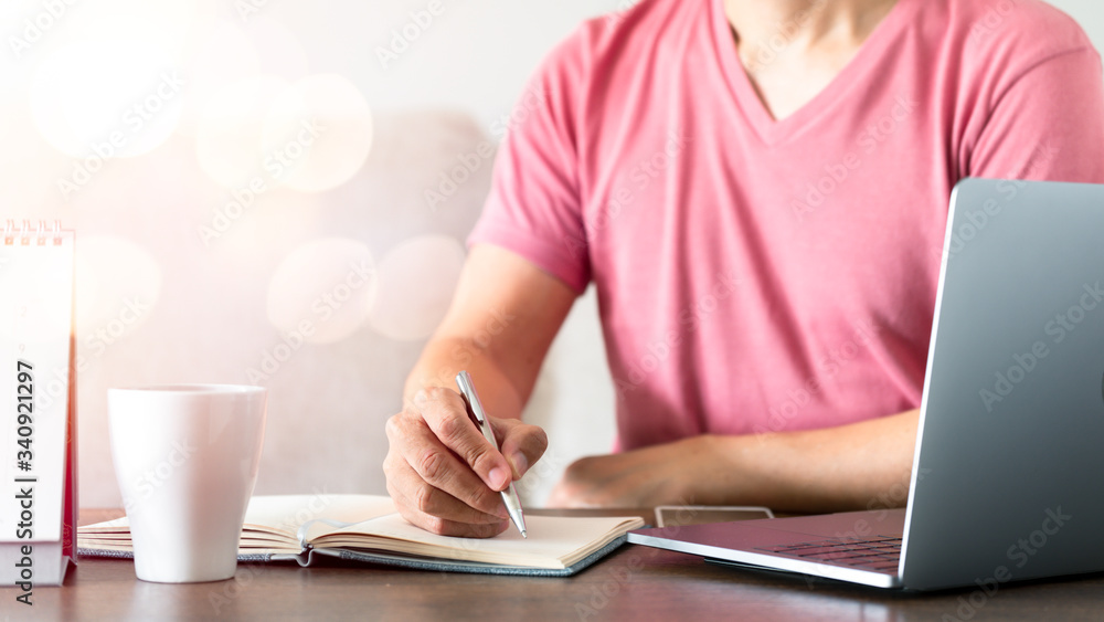 Fototapeta premium Man's hand using pen writing on the notebook on the wood table with technology equipment device while remote working.