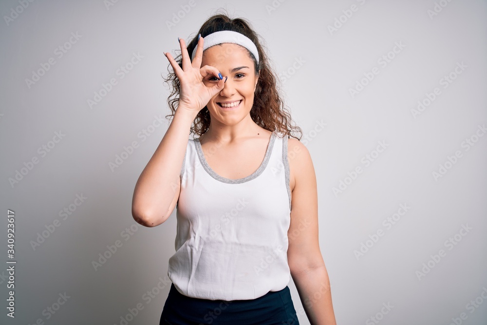 Beautiful sportswoman with curly hair wearing sportswear over isolated white background doing ok gesture with hand smiling, eye looking through fingers with happy face. Beautiful sportswoman with curly hair wearing sportswear over isolated white background doing ok gesture with hand smiling, eye looking through fingers with happy face.