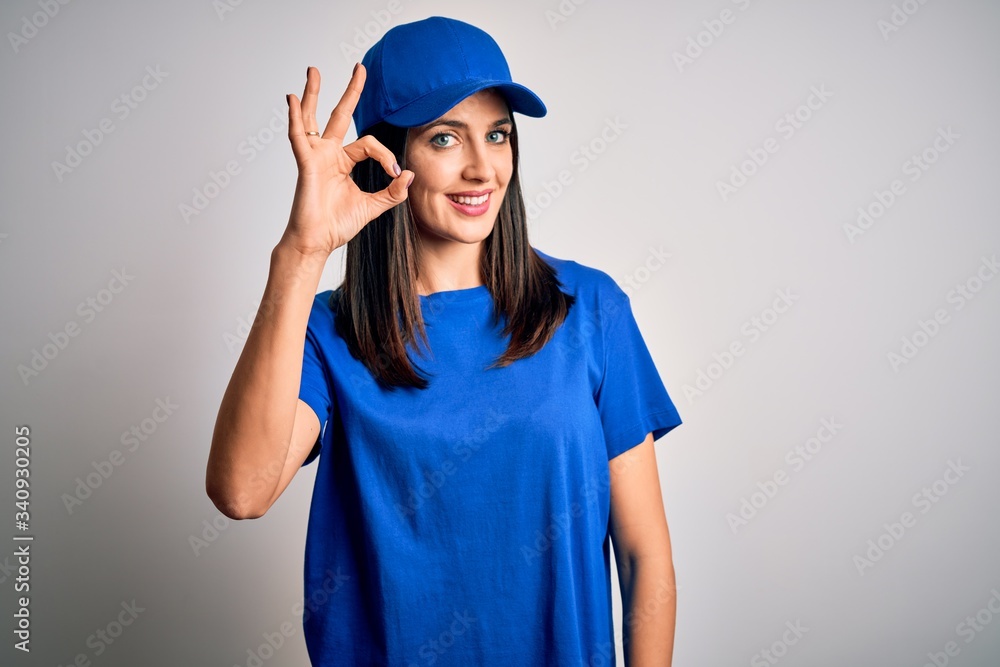 Young delivery woman with blue eyes wearing cap standing over blue background smiling positive doing ok sign with hand and fingers. Successful expression.