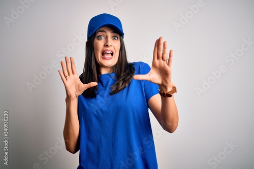 Young delivery woman with blue eyes wearing cap standing over blue background afraid and terrified with fear expression stop gesture with hands, shouting in shock. Panic concept.