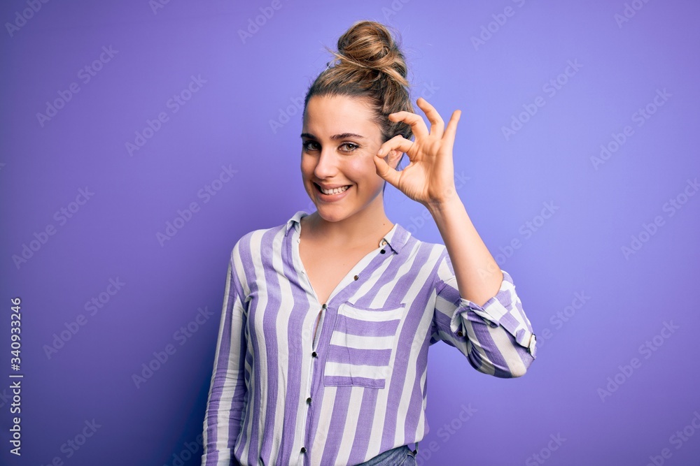 Young beautiful blonde woman wearing casual striped shirt standing over purple background smiling positive doing ok sign with hand and fingers. Successful expression. Young beautiful blonde woman wearing casual striped shirt standing over purple background smiling positive doing ok sign with hand and fingers. Successful expression.
