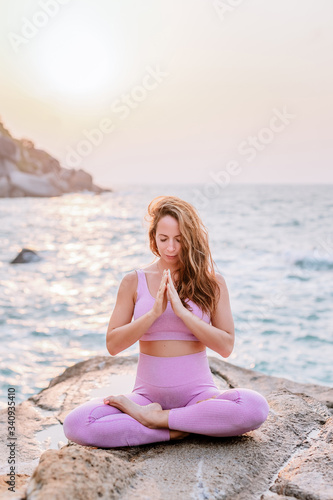Pregnant woman practices yoga, sitting in lotus position on the beach, on the rocks. Hands of girl in a pose meditating. Serenity practicing at sunrise, meditation. Woman in a pink-purple tracksuit.