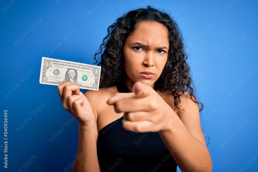 Young beautiful woman with curly hair holding one dollar over isolated blue background pointing with finger to the camera and to you, hand sign, positive and confident gesture from the front Young beautiful woman with curly hair holding one dollar over isolated blue background pointing with finger to the camera and to you, hand sign, positive and confident gesture from the front