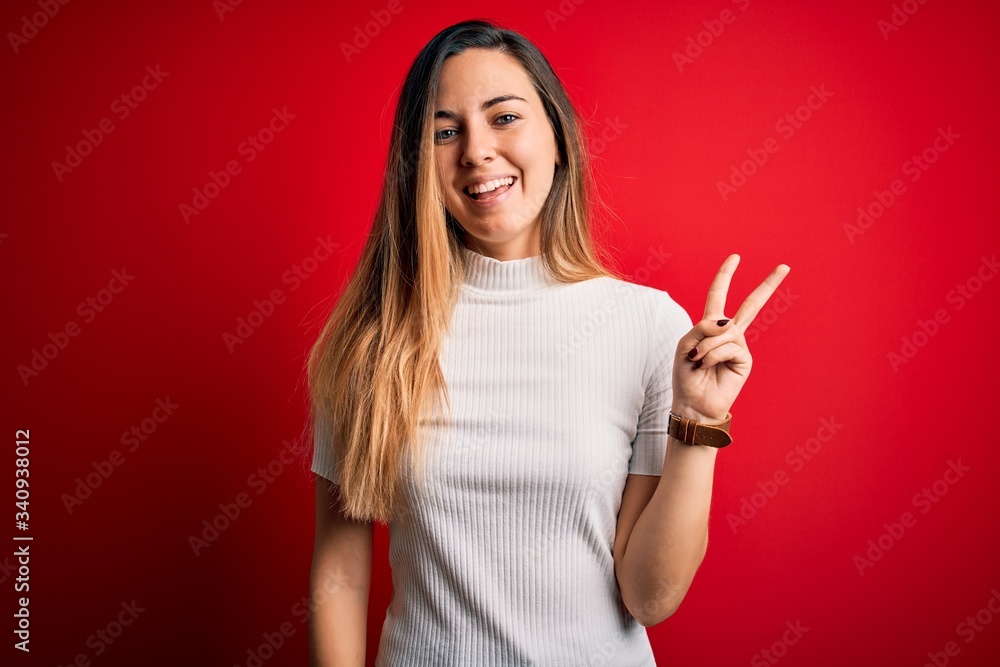 Beautiful blonde woman with blue eyes wearing casual white t-shirt over red background smiling with happy face winking at the camera doing victory sign with fingers. Number two. Beautiful blonde woman with blue eyes wearing casual white t-shirt over red background smiling with happy face winking at the camera doing victory sign with fingers. Number two.
