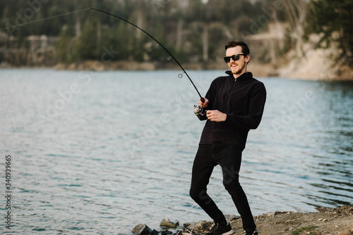 Fisherman with rod, spinning reel on the river bank. Man catching fish, pulling rod while fishing from lake or pond with text space at weekend. Fishing for pike, perch, carp on beach lake or pond.