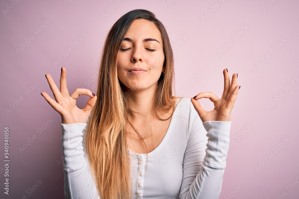 Young beautiful blonde woman with blue eyes wearing white t-shirt over pink background relaxed and smiling with eyes closed doing meditation gesture with fingers. Yoga concept. Young beautiful blonde woman with blue eyes wearing white t-shirt over pink background relaxed and smiling with eyes closed doing meditation gesture with fingers. Yoga concept.