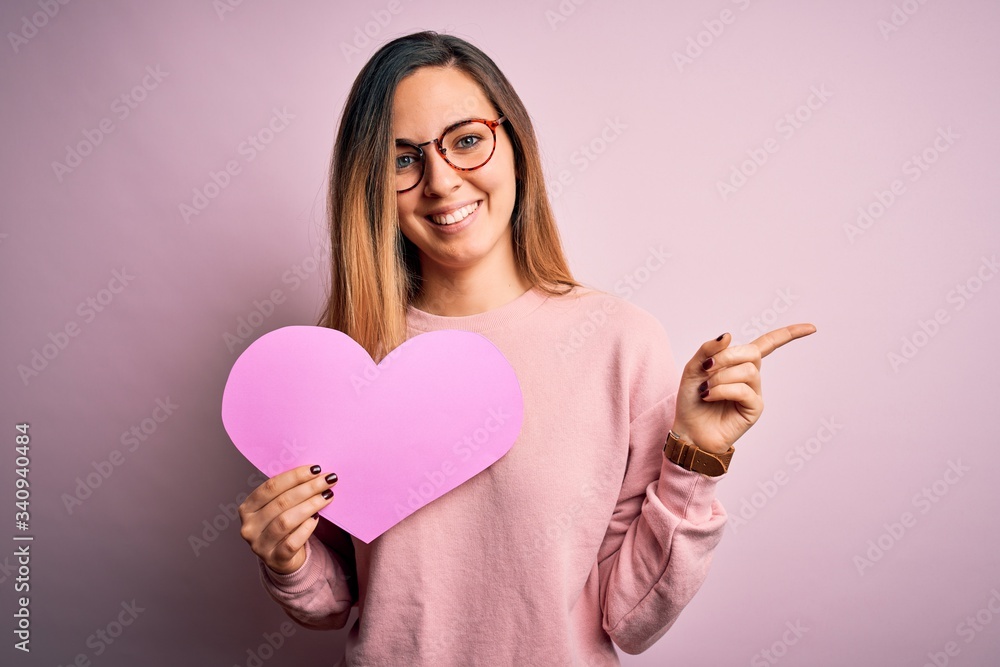Young beautiful blonde romantic woman wearing glasses holding big pink heart very happy pointing with hand and finger to the side Young beautiful blonde romantic woman wearing glasses holding big pink heart very happy pointing with hand and finger to the side