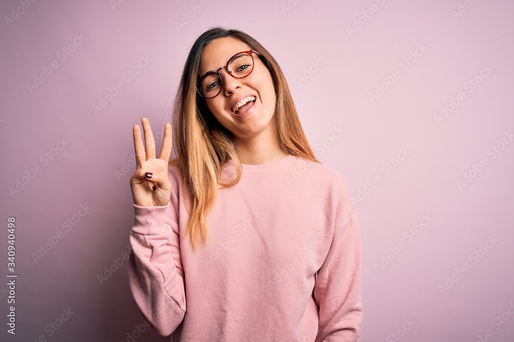 Beautiful blonde woman with blue eyes wearing sweater and glasses over pink background showing and pointing up with fingers number three while smiling confident and happy. Beautiful blonde woman with blue eyes wearing sweater and glasses over pink background showing and pointing up with fingers number three while smiling confident and happy.