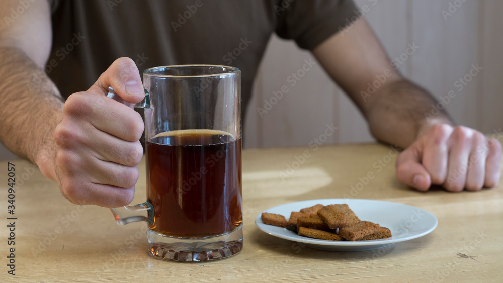 A man sits at a table and holds in his hand a mug of dark beer. On the table is a mug and a plate with bread crumbs. A man sits at a table and holds in his hand a mug of dark beer. On the table is a mug and a plate with bread crumbs.