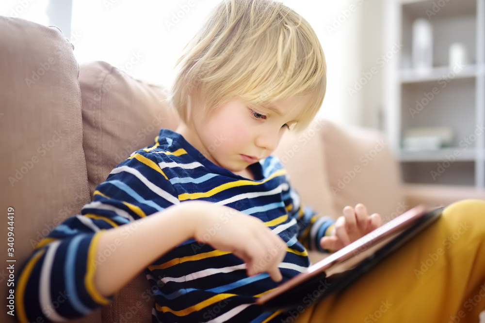Preschooler boy is learning on distance by tablet or playing pc game sitting on couch. Homeschool education and entertainments for kids while coronavirus epidemic. Stay at home during quarantine. Preschooler boy is learning on distance by tablet or playing pc game sitting on couch. Homeschool education and entertainments for kids while coronavirus epidemic. Stay at home during quarantine.