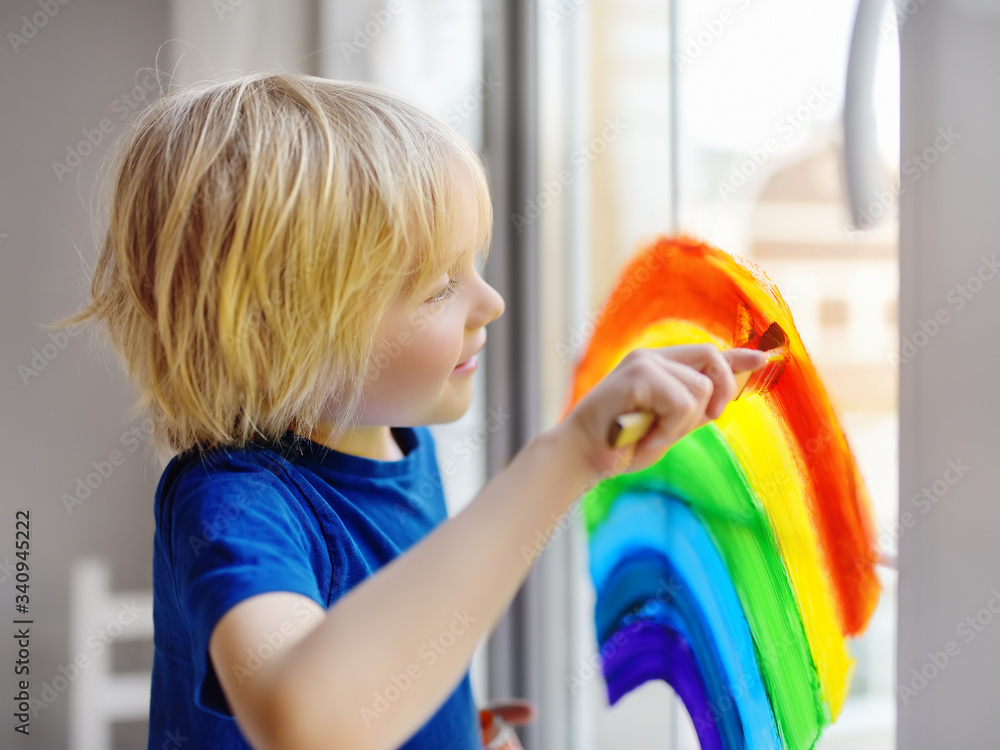 Little boy drawing on window rainbow while coronavirus quarantine. Rainbow sign is symbol of hope, means that everything will be OK. - obrazy, fototapety, plakaty 