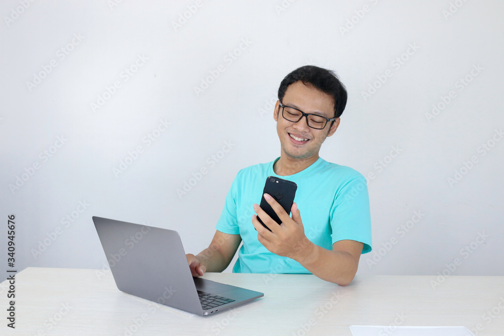 Asian young man happy and smile when he see on smart phone with laptop beside it. Indonesian man wearing blue shirt.