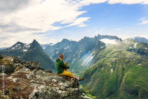 Hiker on the top of norwegian mountain ridge looking to the valley with green hills and river. Norway.